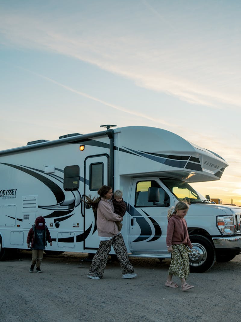 Class C Entegra Coach RV in dirt lot with sun in the background and woman holding baby and two kids walk in front