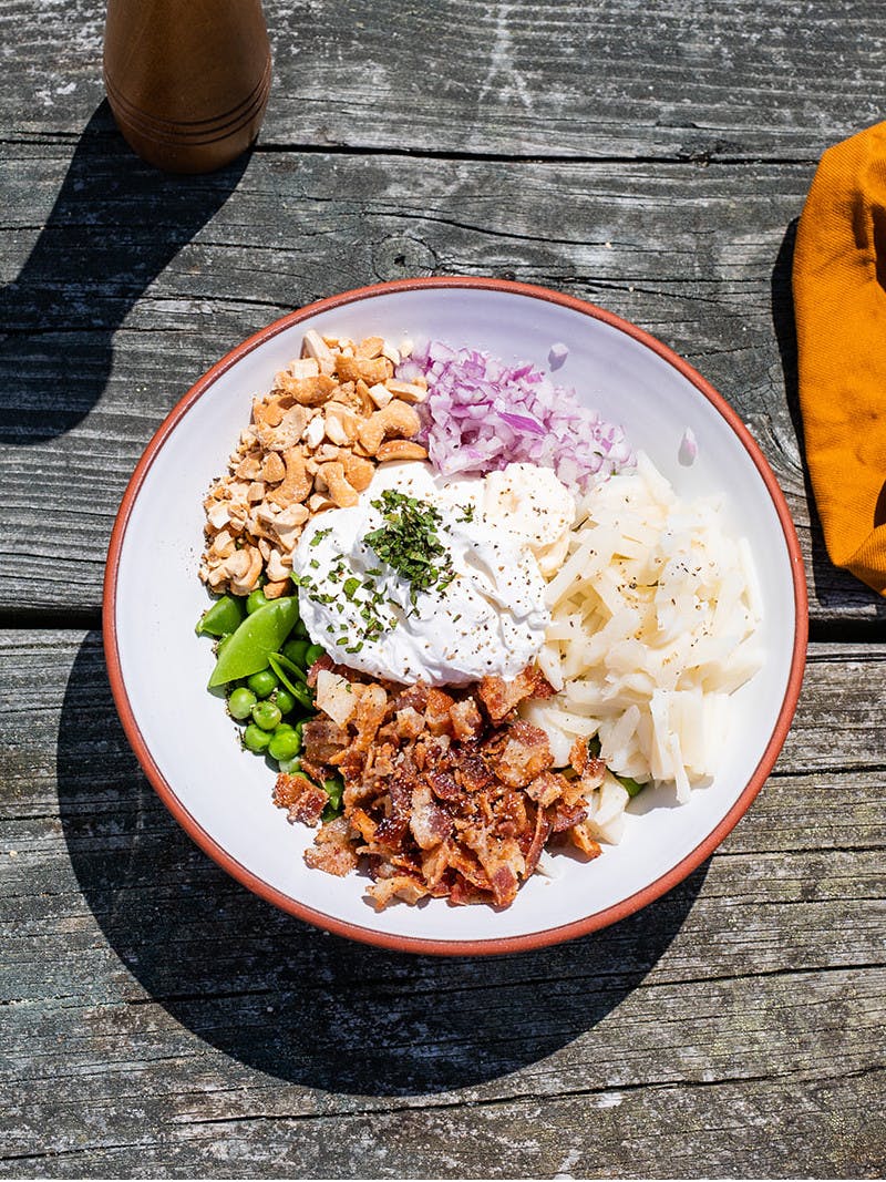 White bowl on picnic table filled with peas, bacon, red onion, water chestnuts, cashews, and sour cream, with two glasses of lemonade.