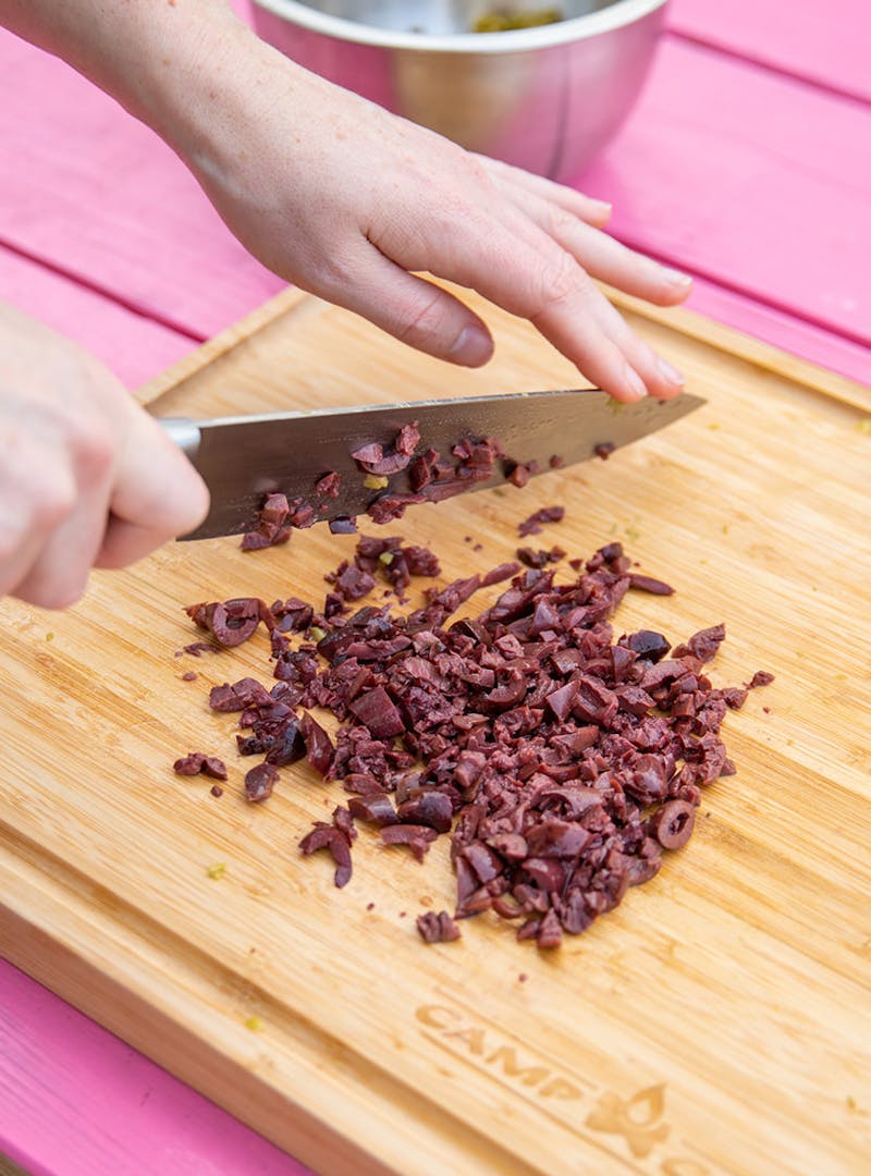 Hands holding a knife above chopped olives on a cutting board.