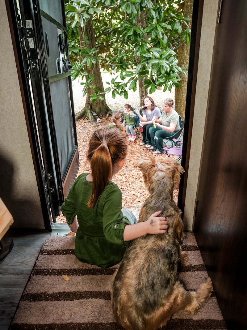 A little girl sits with a dog in an RV doorway, looking outside at her parents and sisters. 