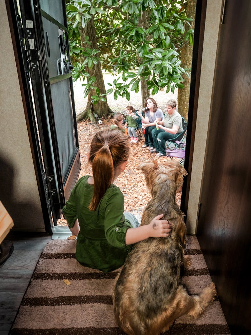 A little girl sits with a dog in an RV doorway, looking outside at her parents and sisters.