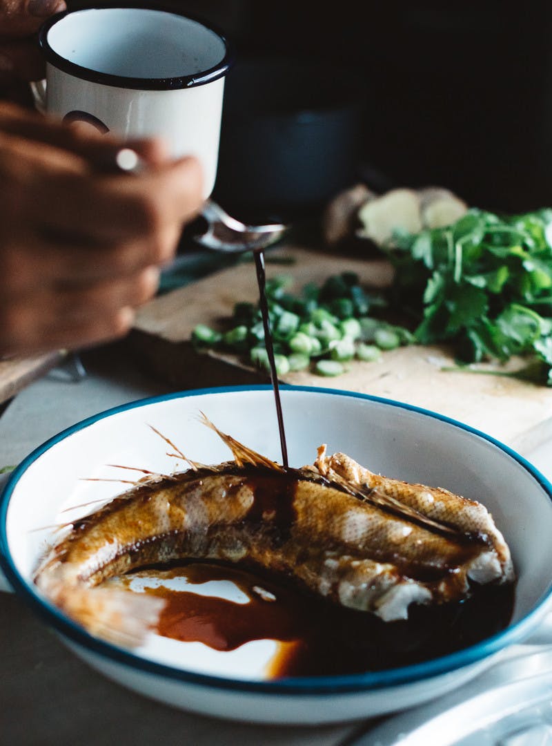 Hand drizzling marinade onto a bowl of cooked fish with cilantro in the background.