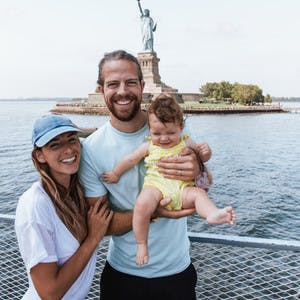A family shot of Bryce Jurgy, his infant daughter and his wife, standing on a boat with the Statue of Liberty in the background.