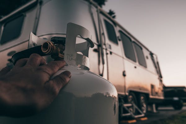 A close up shot of a hand on a propane tank with the sun setting on an RV in the background. 