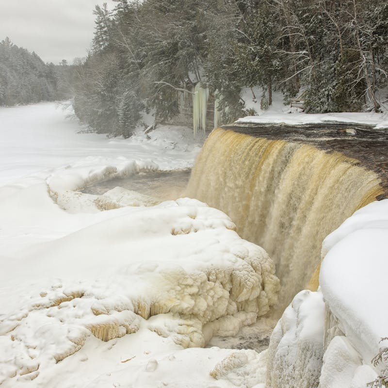 Wide waterfall with brownish colored water falling into a frozen and snow covered river, surrounded by snow