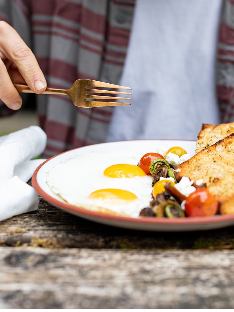 A plate of breakfast food, including eggs, toast, and vegetables, in front of a man holding a fork, about to break open the sunny-side eggs.