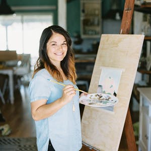 Coley Kuyper stands smiling in front of an easel, in a blue shirt, holding a paint brush and palette, painting a picture.