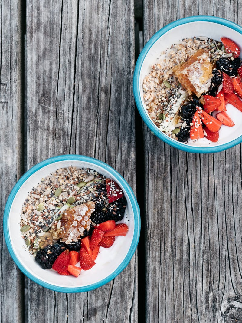White bowls with a sky-blue rim sitting on a gray, aged wood picnic table. The bowls hold servings of overnight oats topped with colorful stripes of berries, chia seeds and honeycomb.