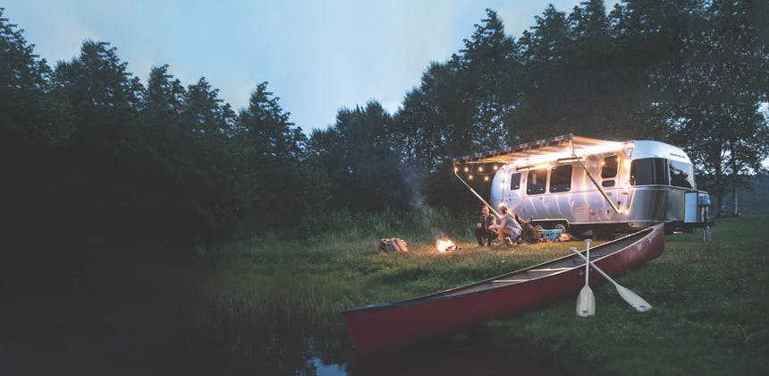Airstream travel trailer RV on edge of lake with canoe, young couple sitting by camp fire with dog