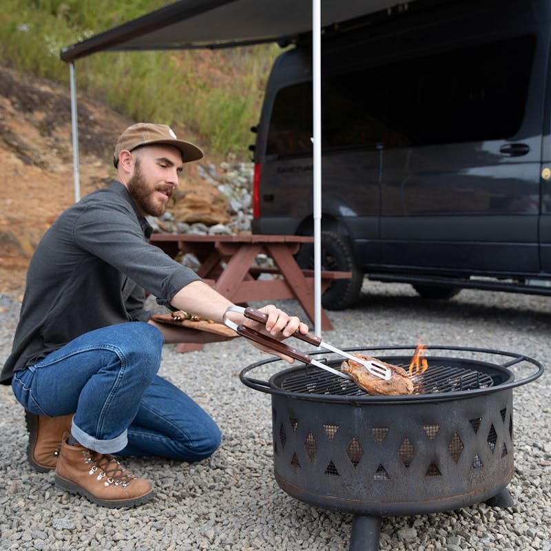 A man cooking Cornish hens over an open flame at a campsite.