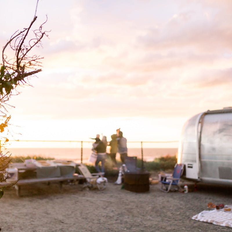 A wide shot of the Thrane family watching the sun set by a fence next to an Airstream Travel Trailer RV, with the beach & ocean in the background.