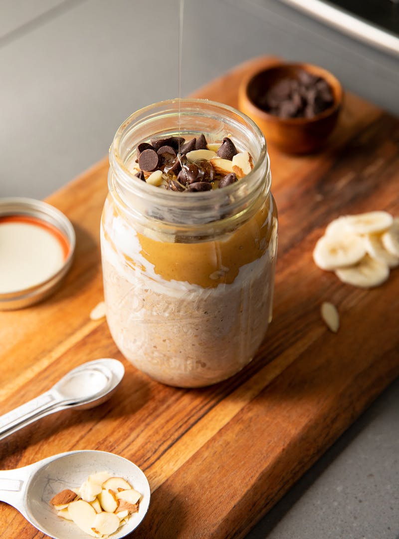 Honey drizzling into a mason jar with overnight oats topped with chocolate chips, peanut butter and almonds.