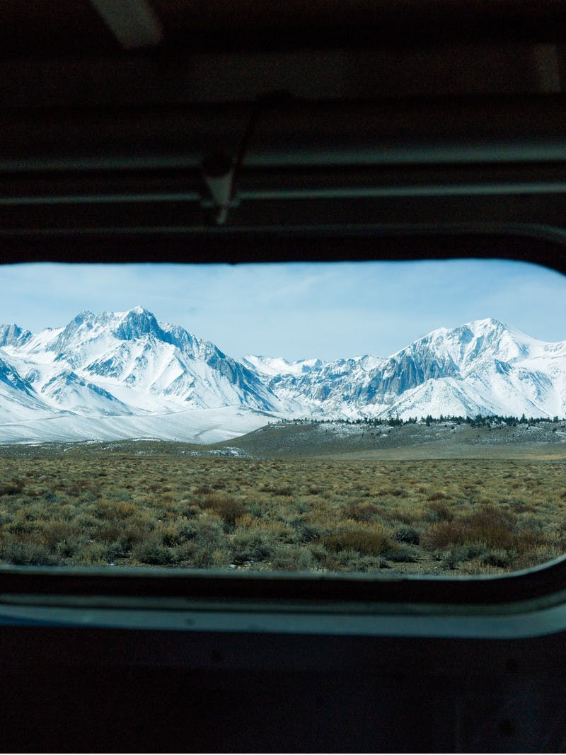 Snowy mountain peaks in the distance, seen through an oblong, rounded RV window. 