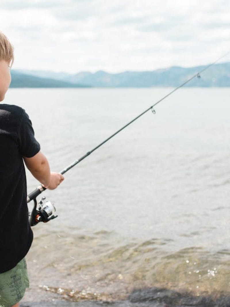 Young boy in black t-shirt holds fishing pole next to lake