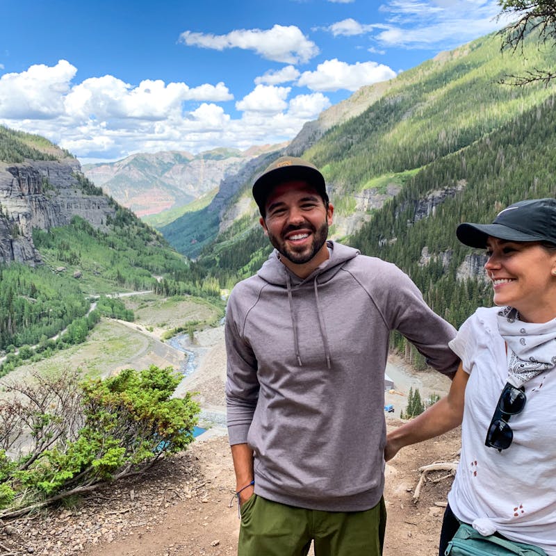 Jason and Dawn smiling for a photo on a ledge in a valley of mountains and trees.