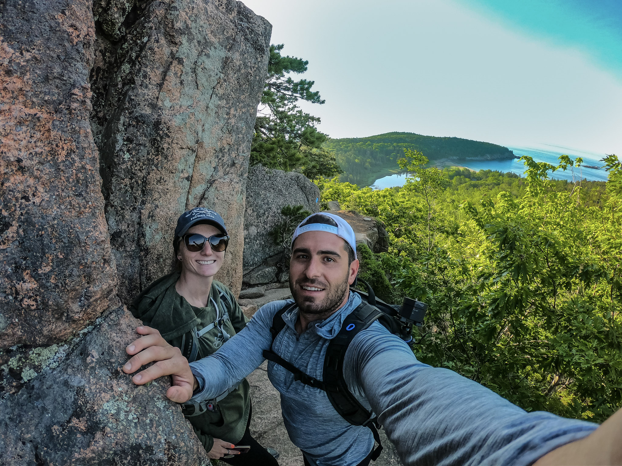 Jesse and Mel taking a selfie from the side of a rocky cliff, with a panoramic landscape in the background. 