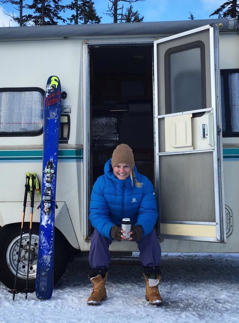 Blonde woman with hair in a braid wearing a hat, bundled up in a blue winter jacket with pants and boots, sits in her RV door, holding a drink, and smiling at the camera. Her skis and ski sticks sit against her RV next to her.