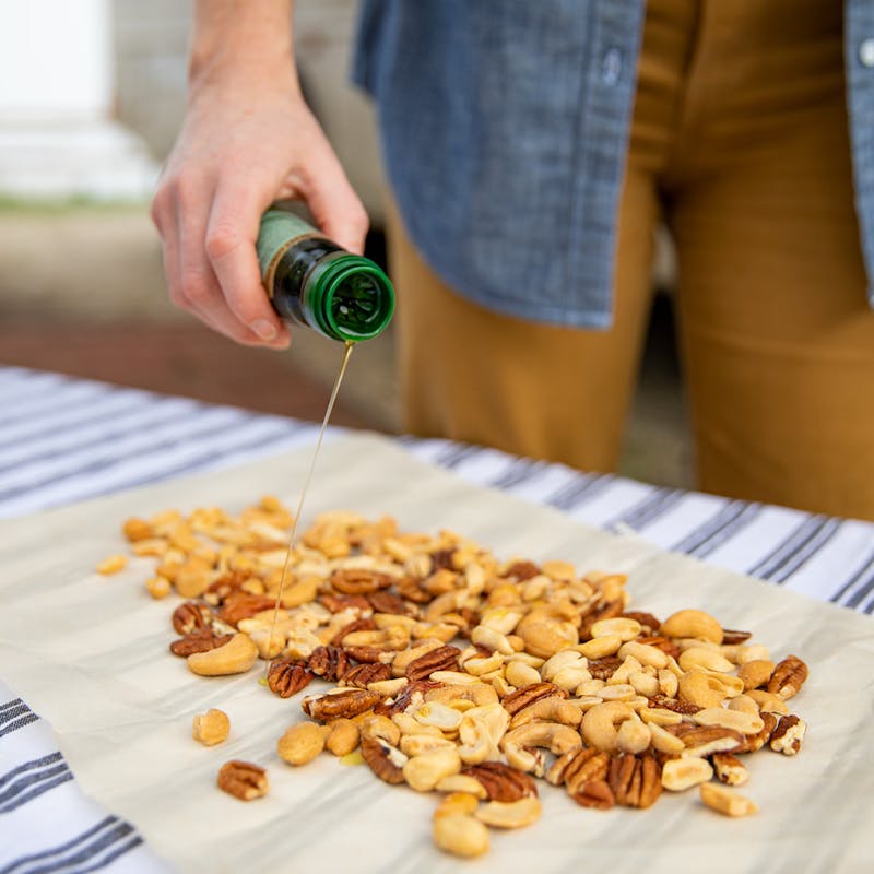 A hand drizzling oil over mixed nuts spread on parchment paper.