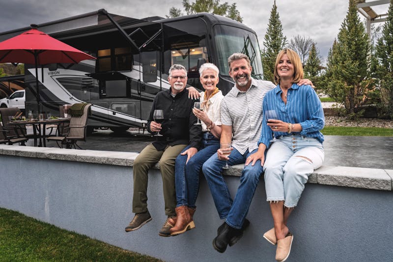 Two couples sit on a ledge drinking wine next to a campsite with an Entegra vision motorhome