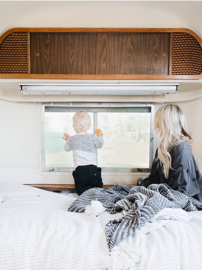 A blonde woman and a small blonde child snuggled in blankets in an RV bed, looking out the window at a beautiful view.