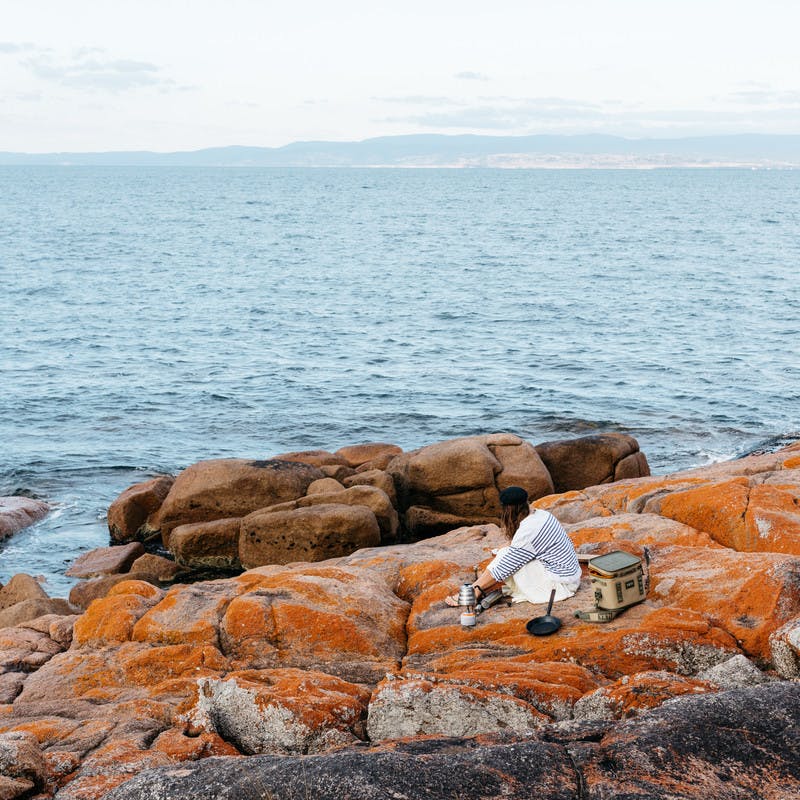 Sarah Glover from a distance, sitting on orange rocks with the ocean, preparing breakfast.