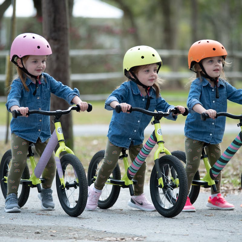 Triplet little girls riding bikes with color coded helmets on their heads.