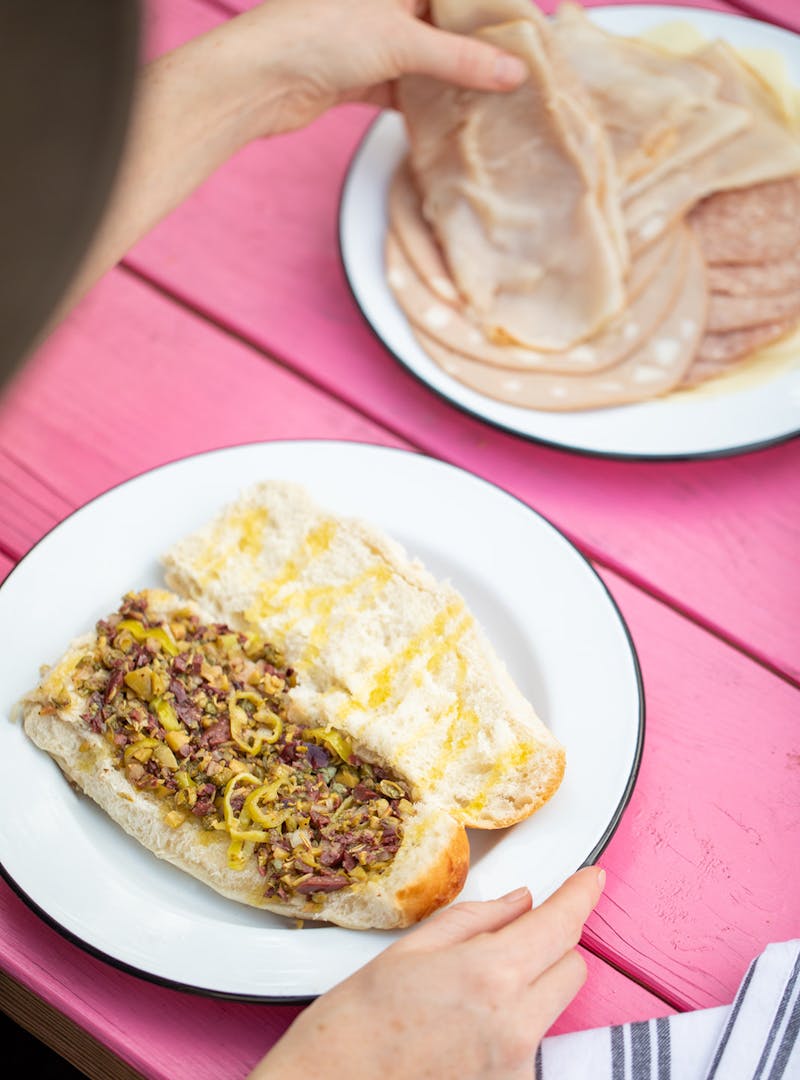 A hand grabbing lunchmeat to layer on a roll already dressed with olive tapenade.