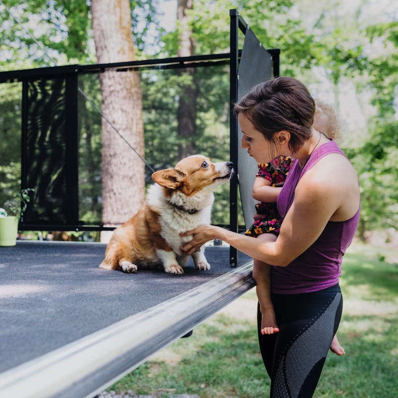 A woman holding her daughter and giving a corgi on an RV porch a kiss.