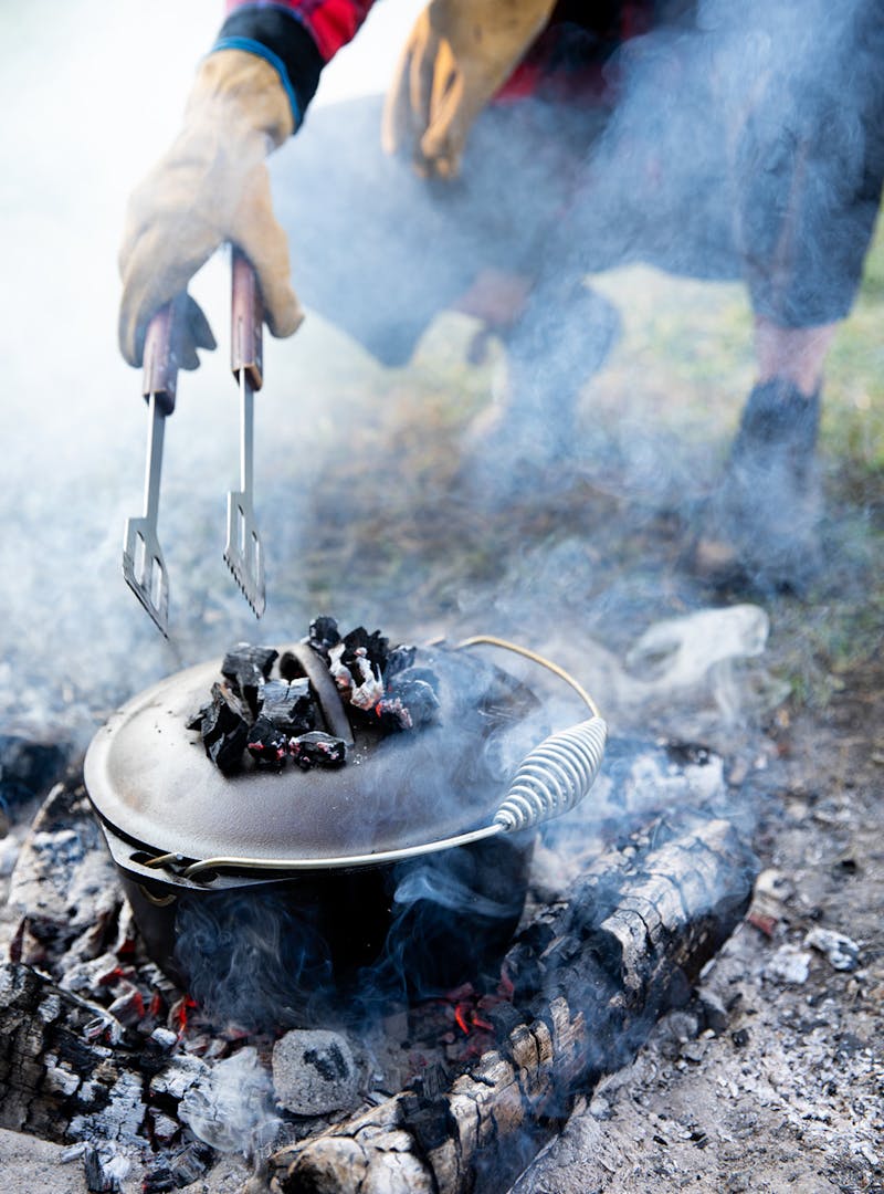 A dutch oven cooks over an open fire with coals on top of the lid.