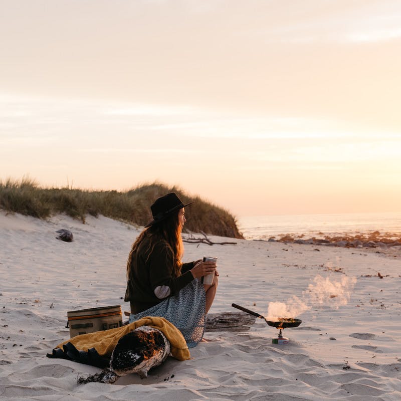 A woman cooking over a small fire on the beach at sunrise.