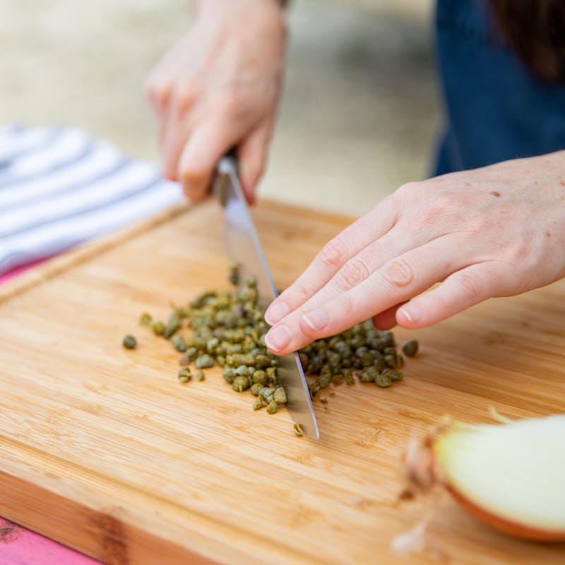 Hands holding a knife that chops capers on a wooden cutting board.