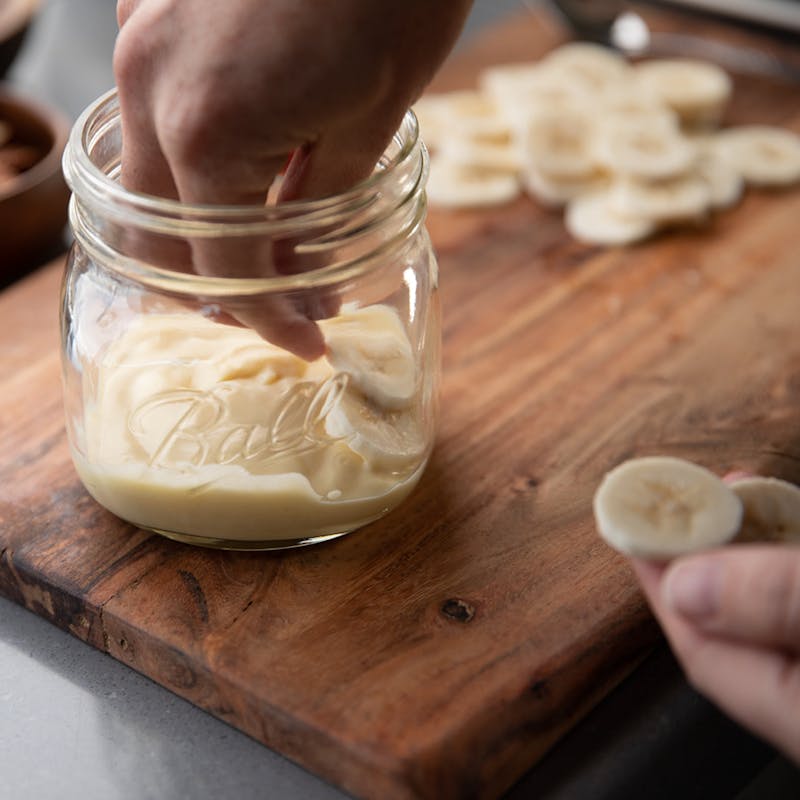 A hand layering banana slices on top of a layer of pudding in a glass jar.