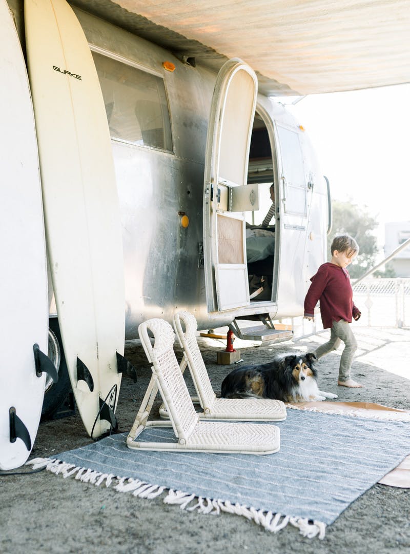 Child leaving through the front door of the Airstream RV, two surfboards rest against the side, with two beach chairs, and a mat on the ground.