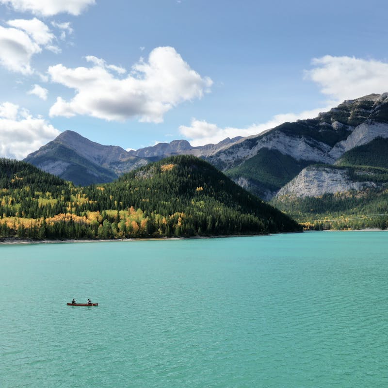 An overhead shot of a tiny canoe on still, turquoise waters, with mountains looming in the background and trees changing color.