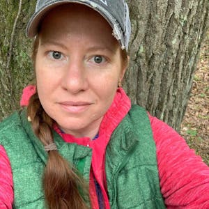A selfie of Amy Lundquist-Stachowiak sitting in front of a tree wearing a hat and green vest