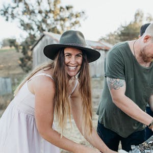 Sarah Glover, in a white sun dress and a felt hat, cooking over grill and smiling at the camera.