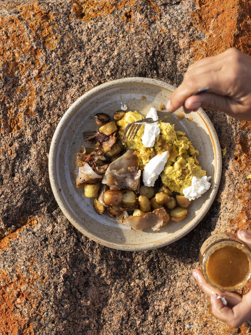 Hand holding a fork over a bowl of eggs, potatoes, and sausage, resting on an orange rock.