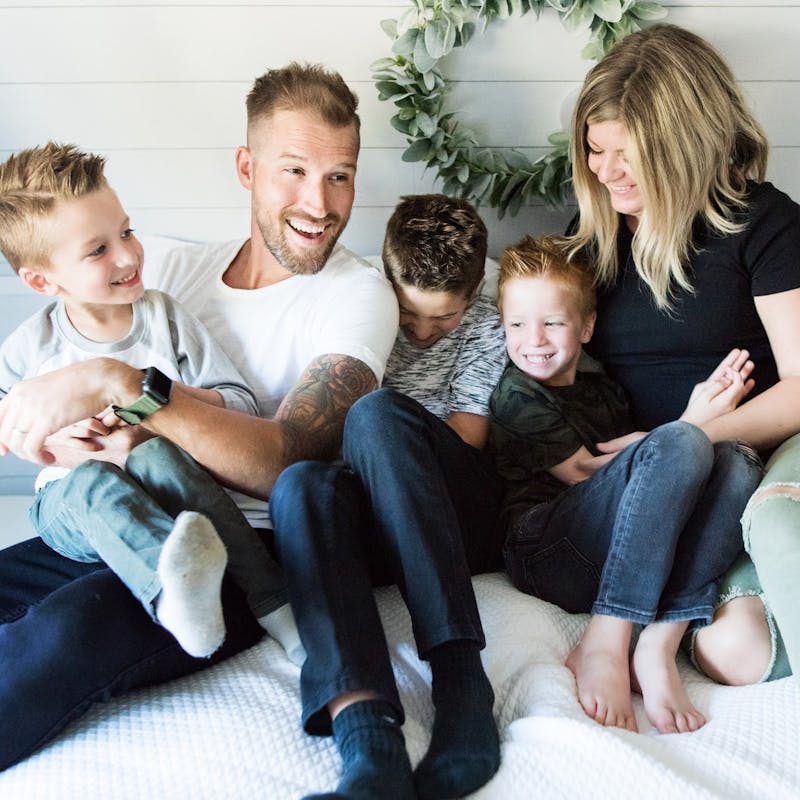 Two parents and three little boys cuddled together on a bed.
