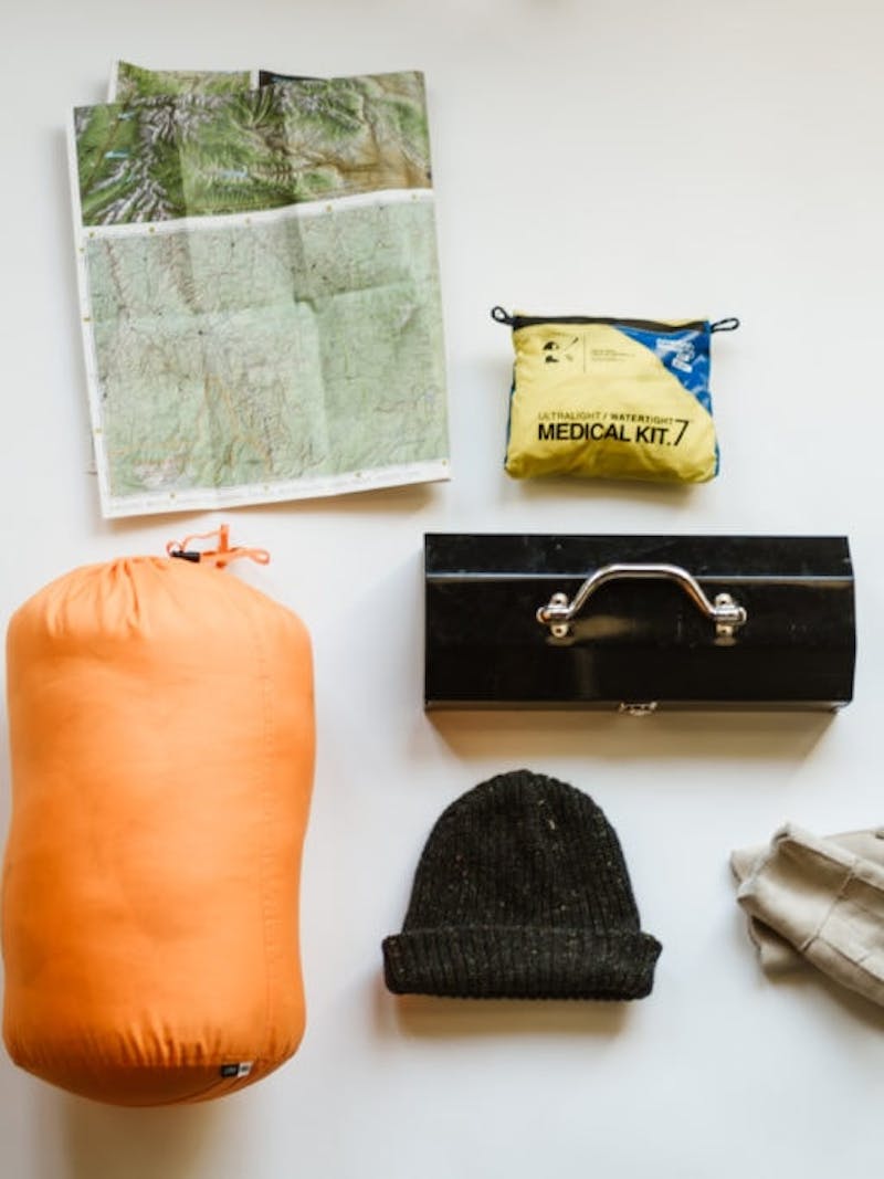 Camping items laid out on white background, including sleeping bag, map, medical kit, hat, tool box, and gloves
