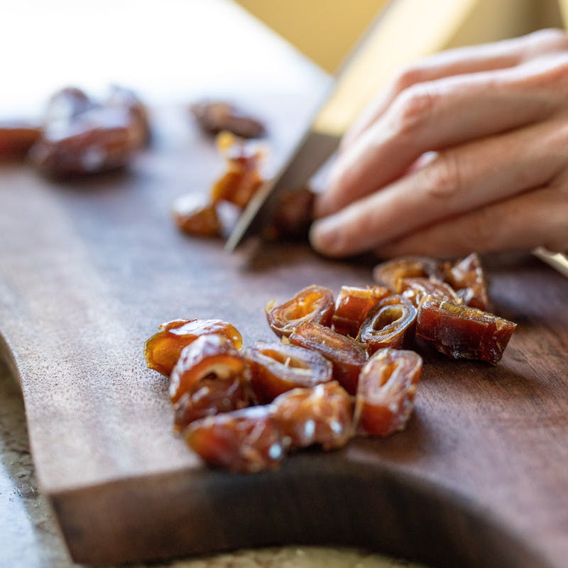 Close up of hands slicing dried dates with a knife on a cutting board.