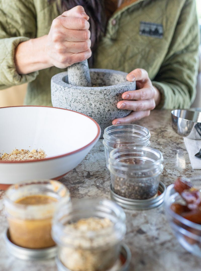 Woman using a mortar and pestle to grind hazelnuts, with mason jars and a white bowl.