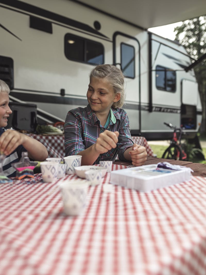 two kids sit at a picnic table at a campsite by an RV