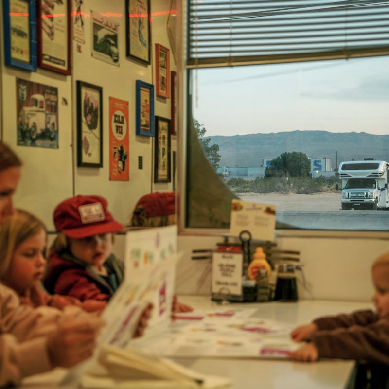 Mom sits at a diner table with three young kids reading menus while class c motorhome sits outside window