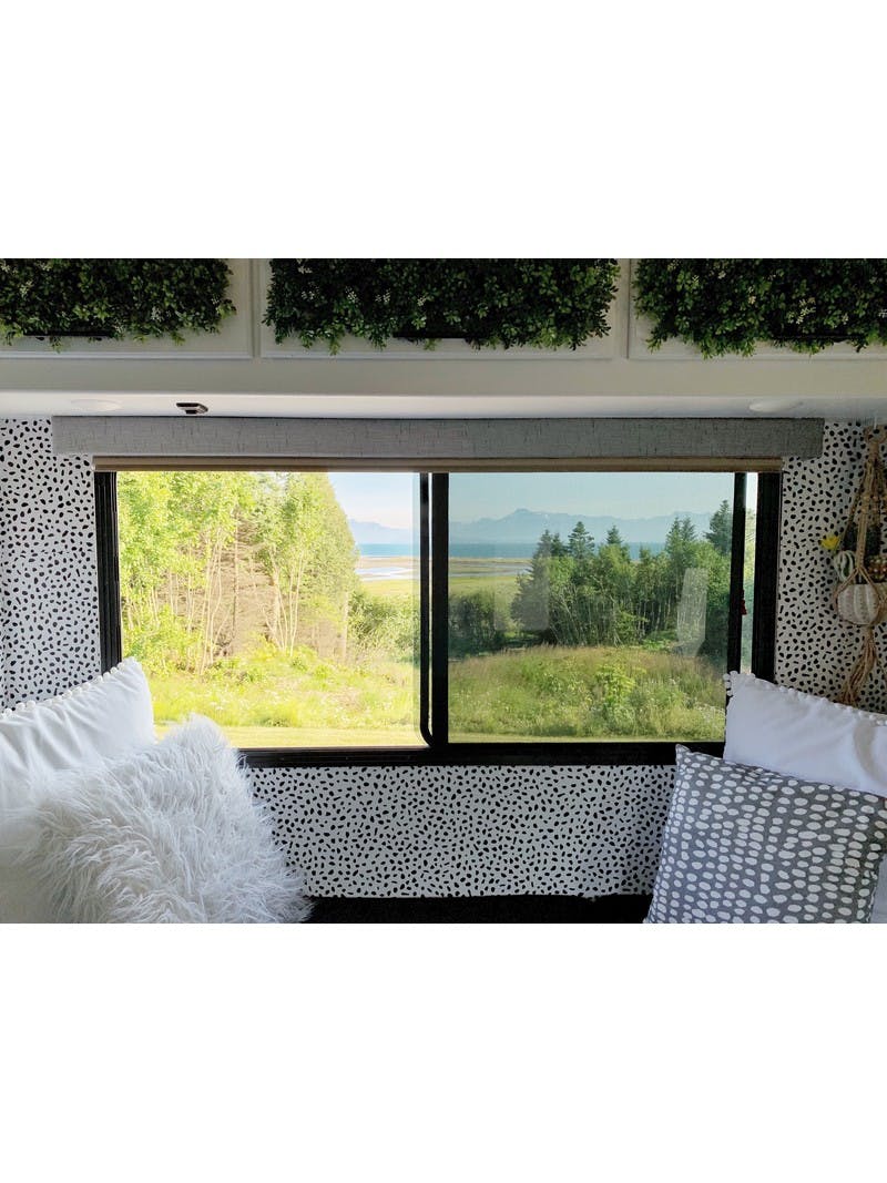 A view from the inside of an RV with comfortable pillows, polka dot wallpaper and a large window looking out to green trees and mountains in Homer, Alaska.
