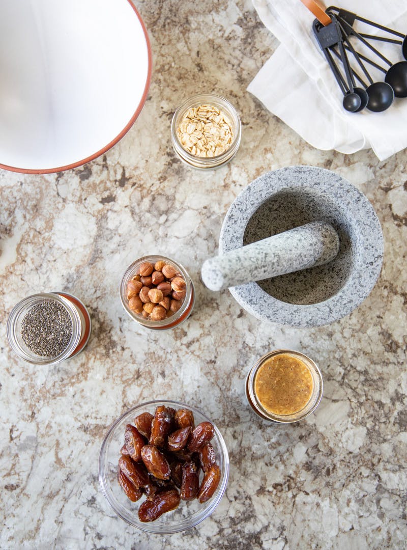 Various bowls and jars on a granite countertop, includes dates, chia seeds, hazelnuts, oats and nut butter.