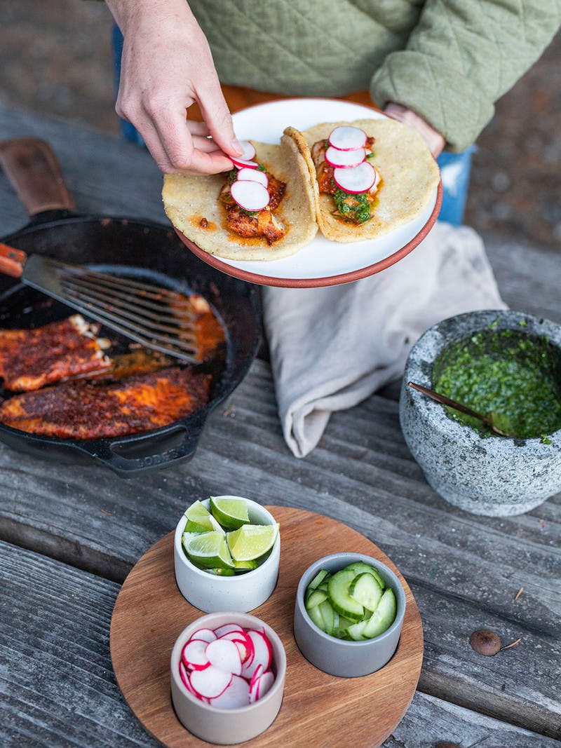 Two blackened rockfish tacos with traditional chimichurri and fire roasted tortillas on a plate, while man tops tacos with radishes.