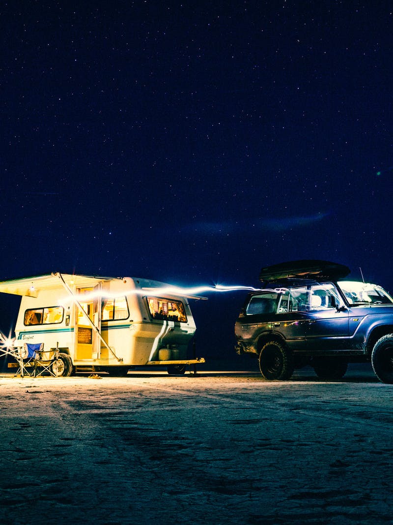 Dark photo with a blue car in front of a lit up trailer, with the awning out, door open, and chairs set up outside the trailer.