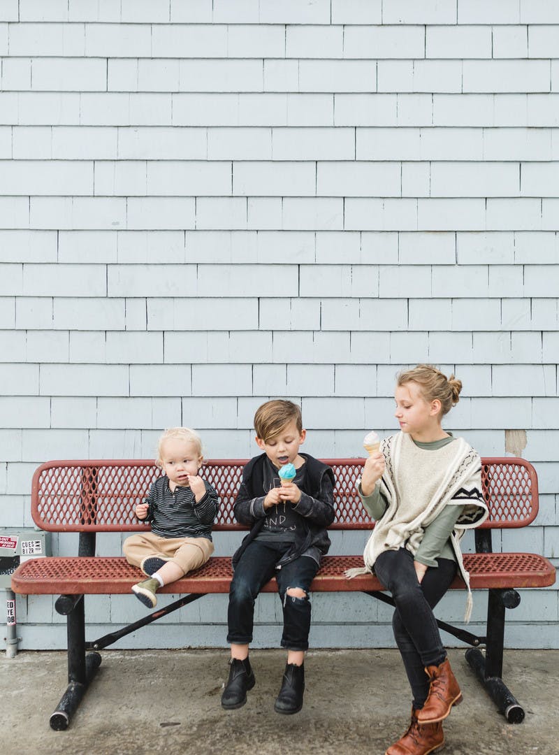 Three kids sit on a red bench eating ice cream cones.