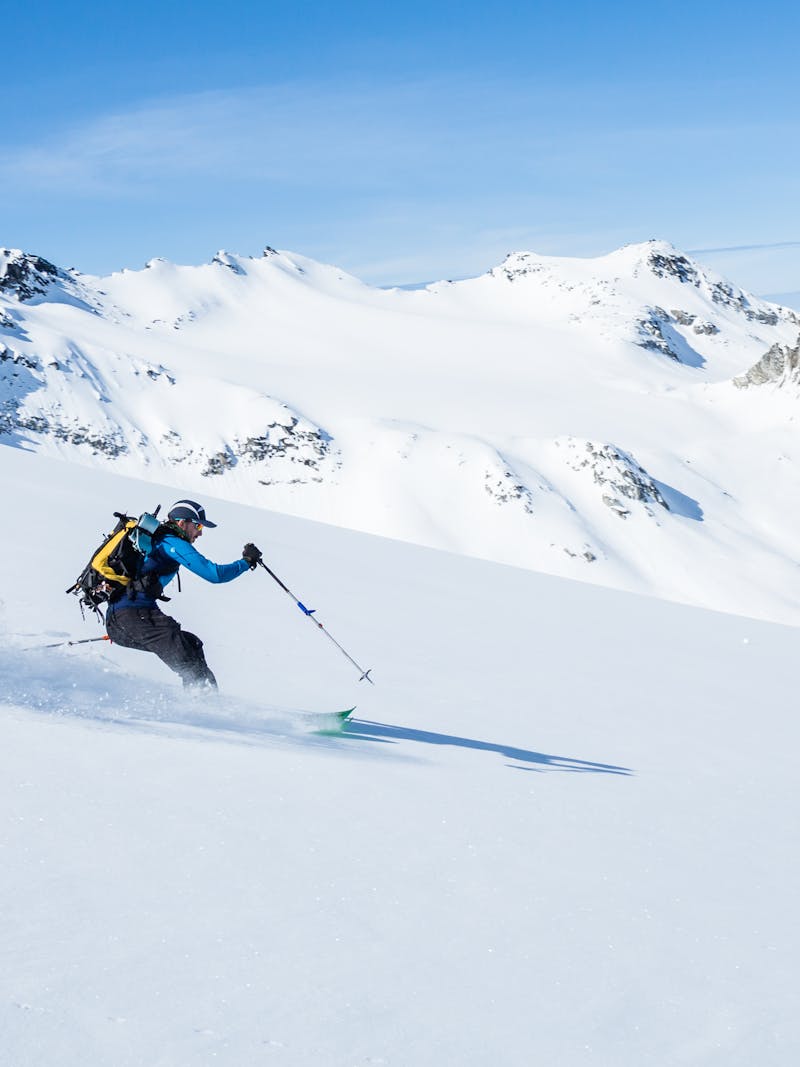 Man with yellow backpack skis down snowy mountain with jagged mountain peaks in the background