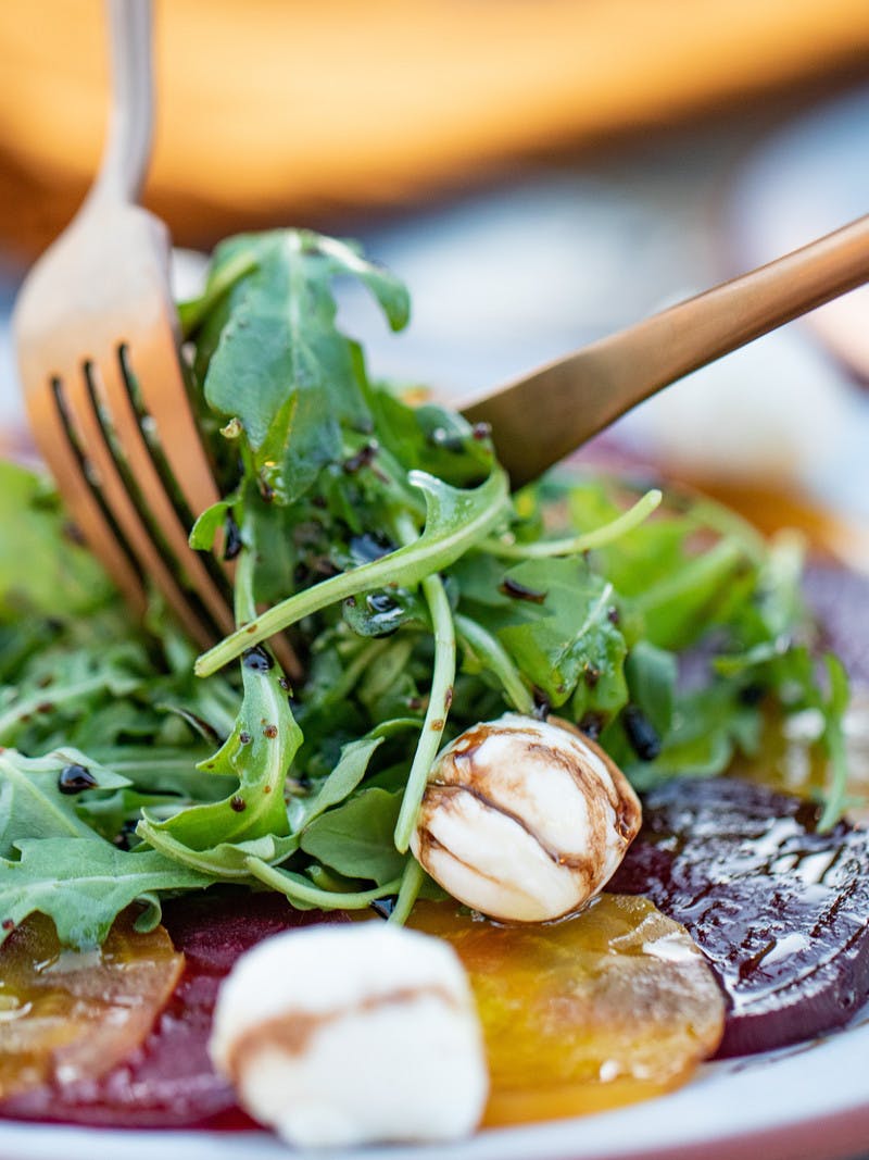 Close up of gold and red beats, topped with arugula, mozzarella cheese balls, and drizzled with a balsamic dressing, includes a knife and fork.